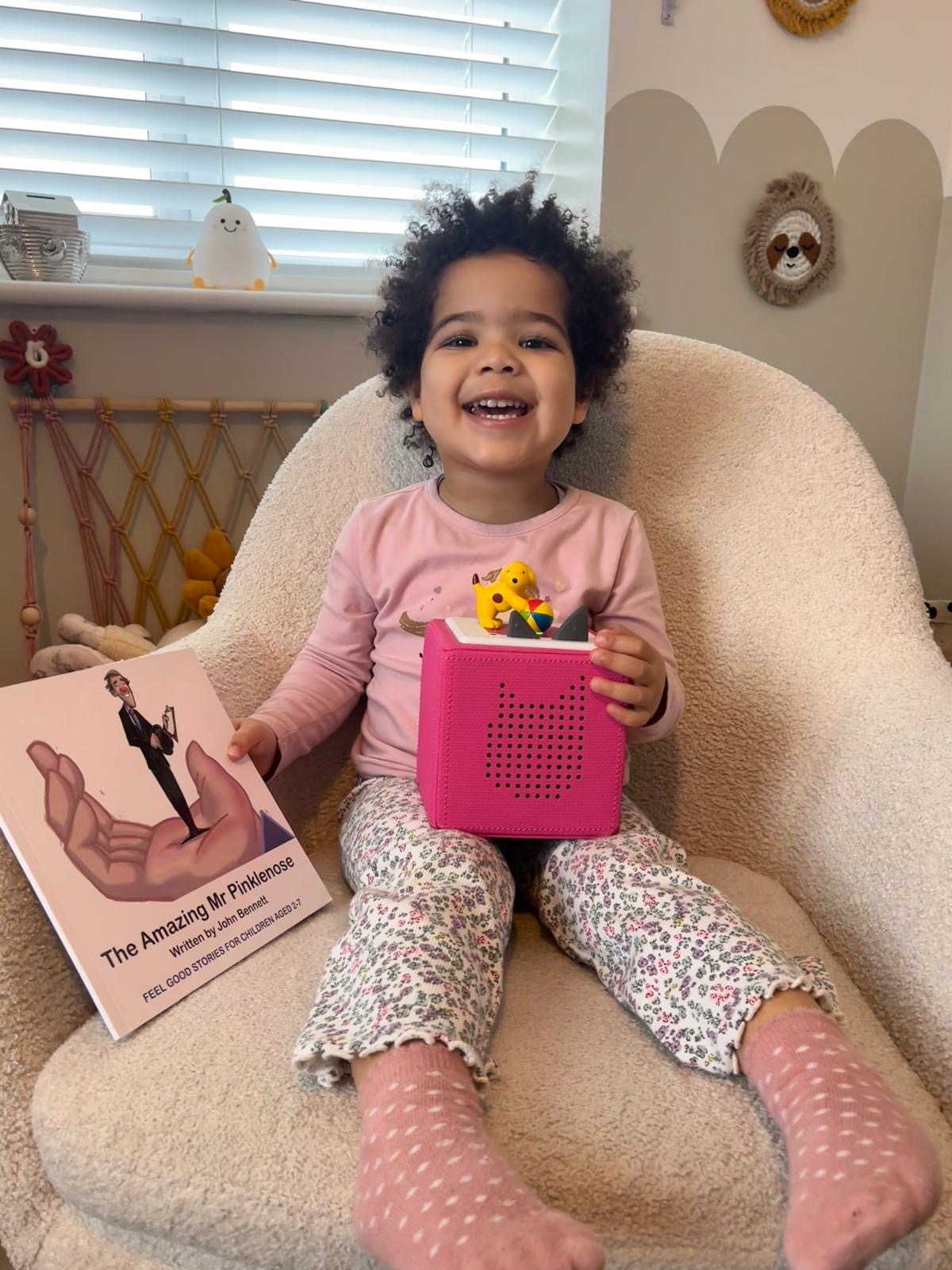 A young girl is sat with her Mr Pinklenose book and her Tonie box that she uses to listen to her granddad reading his stories.
