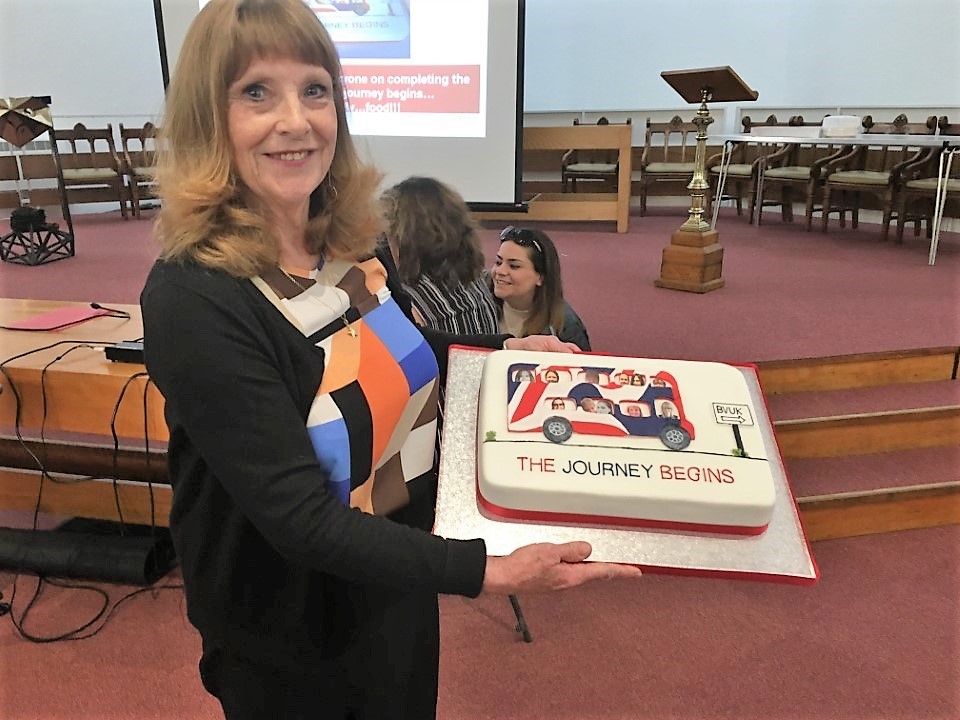 Jennie looks into camera and is holding a cake with a bus on it in the Blind Veterans UK colours. The caption below the bus reads 'the journey begins'.