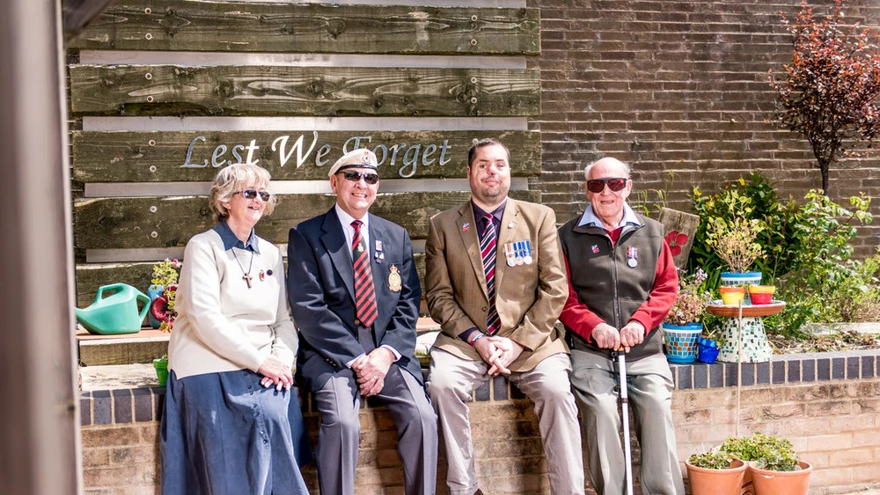 A group of blind veterans seated on a wall, smiling in front of a sign that reads "Lest We Forget"