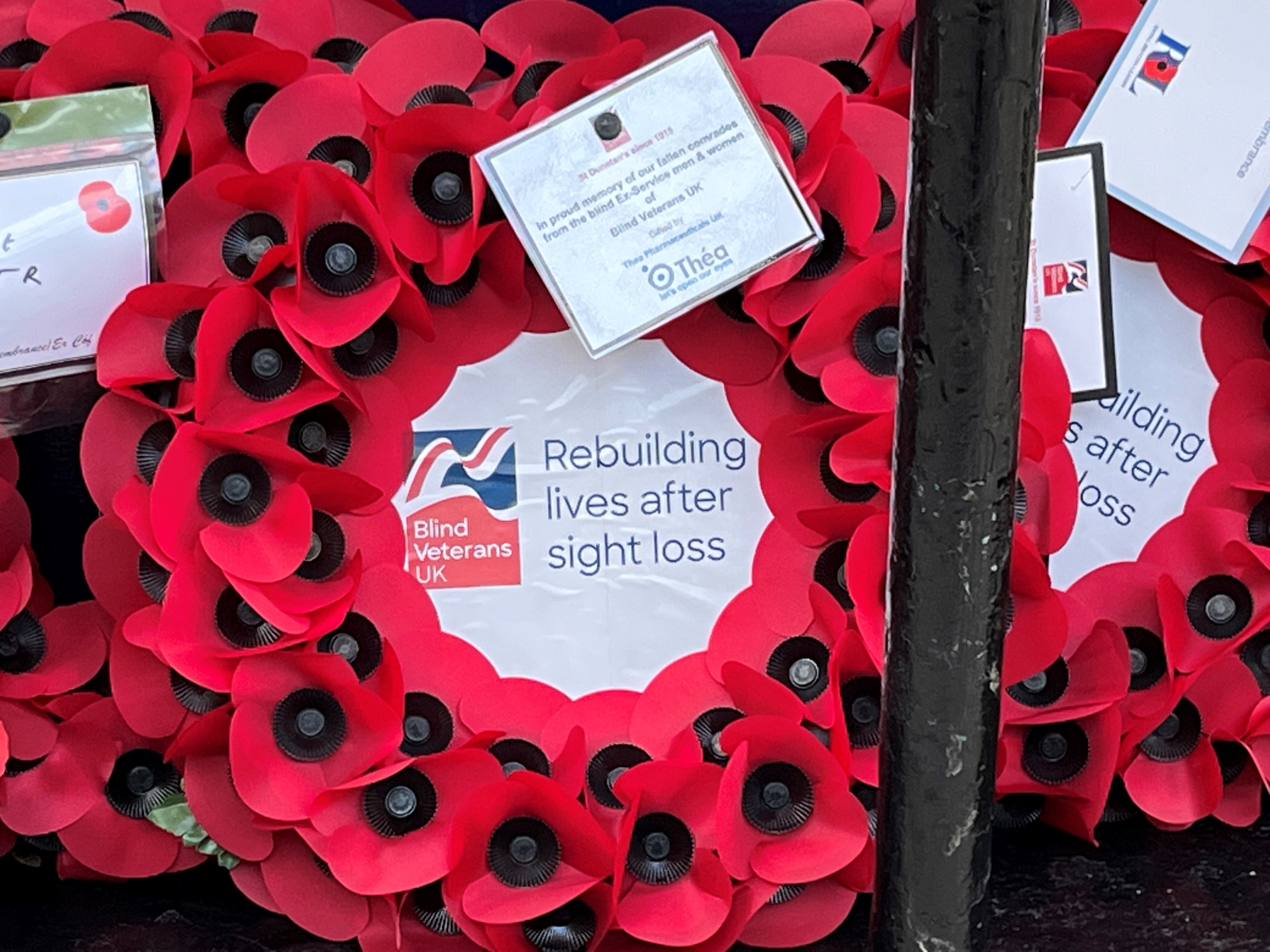 Close up of poppy wreath with Blind Veterans UK logo in the centre and a label attached which reads "In proud memory of our fallen comrades from the blind ex-Service men and women of Blind Veterans UK gifted by Thea Pharmaceuticals