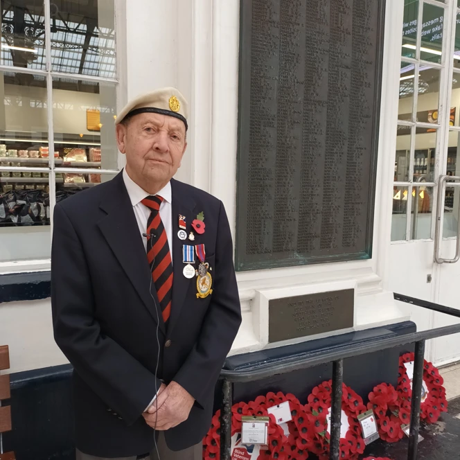 Tony wearing his medals and stood in front of Brighton Train Station War Memorial which has a number of poppy wreaths laid against it