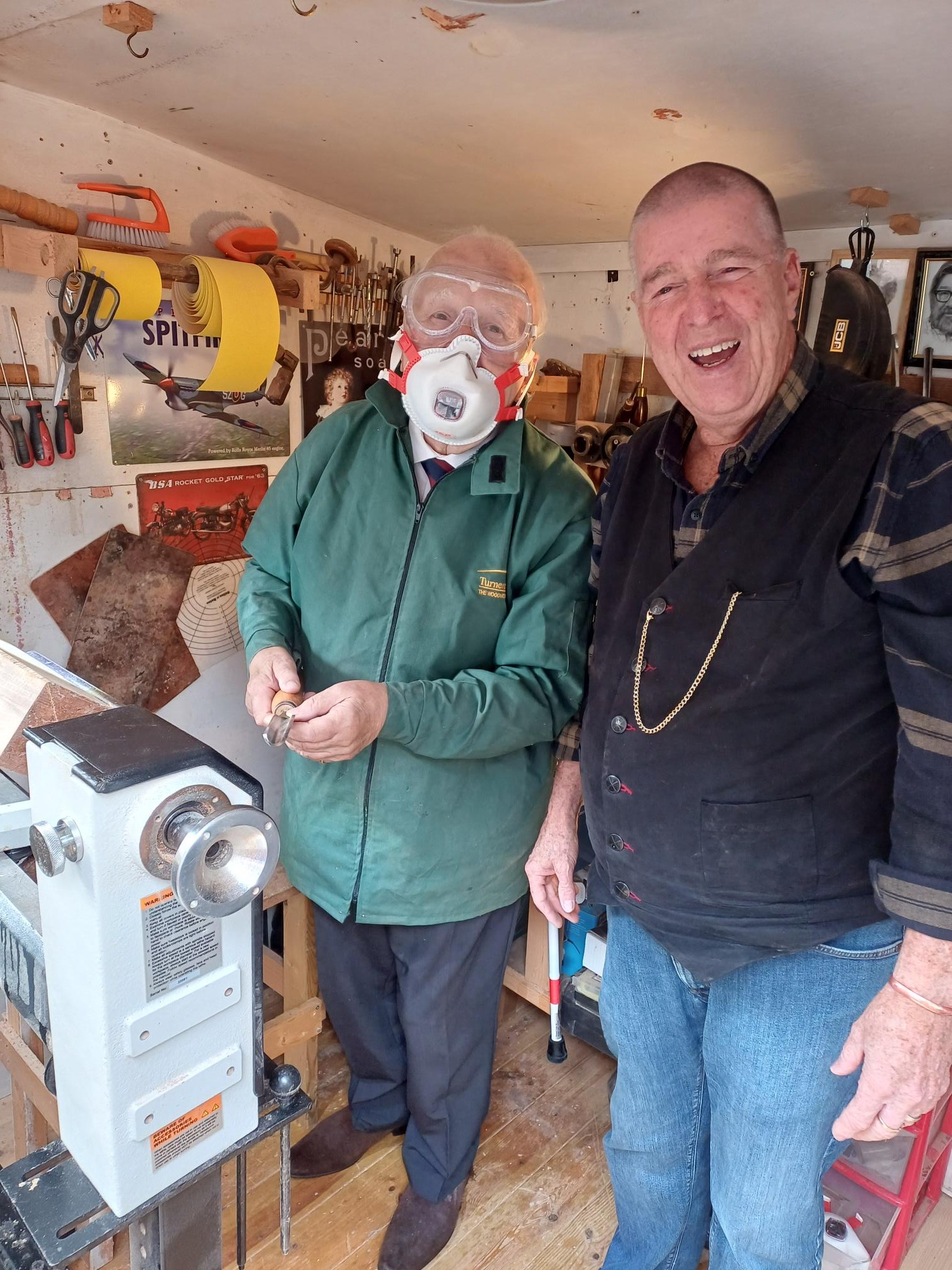 Brian wears a mask and stands in front of carpentry machinery with Kevin standing next to him smiling