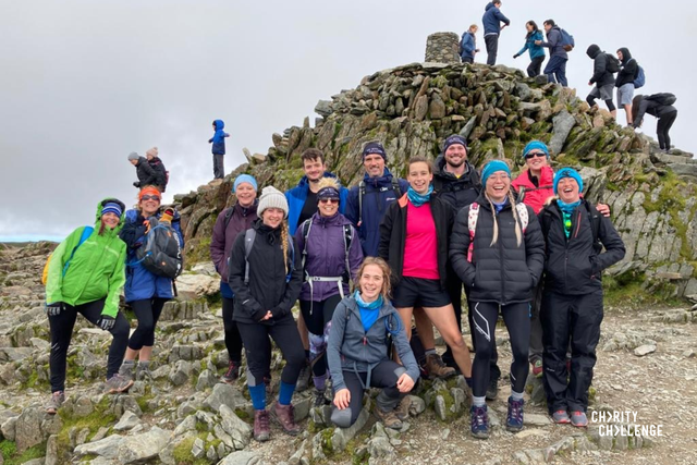 A group of challengers wearing walking gear, smiling widely at the camera.  They are on a plateau of rock just in front of the pile of stones upon which sites the toposcope that marks the peak of Snowdon.