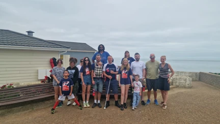 A group photo of Ian with his white cane in the centre surrounded by 13 of his family and friends.