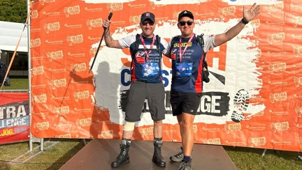 Charlie and Rich stood on a stage with a South Coast Ultra backdrop. The pair are in their Blind Veterans UK t-shirts, shorts and walking boots and have one hand each in the air