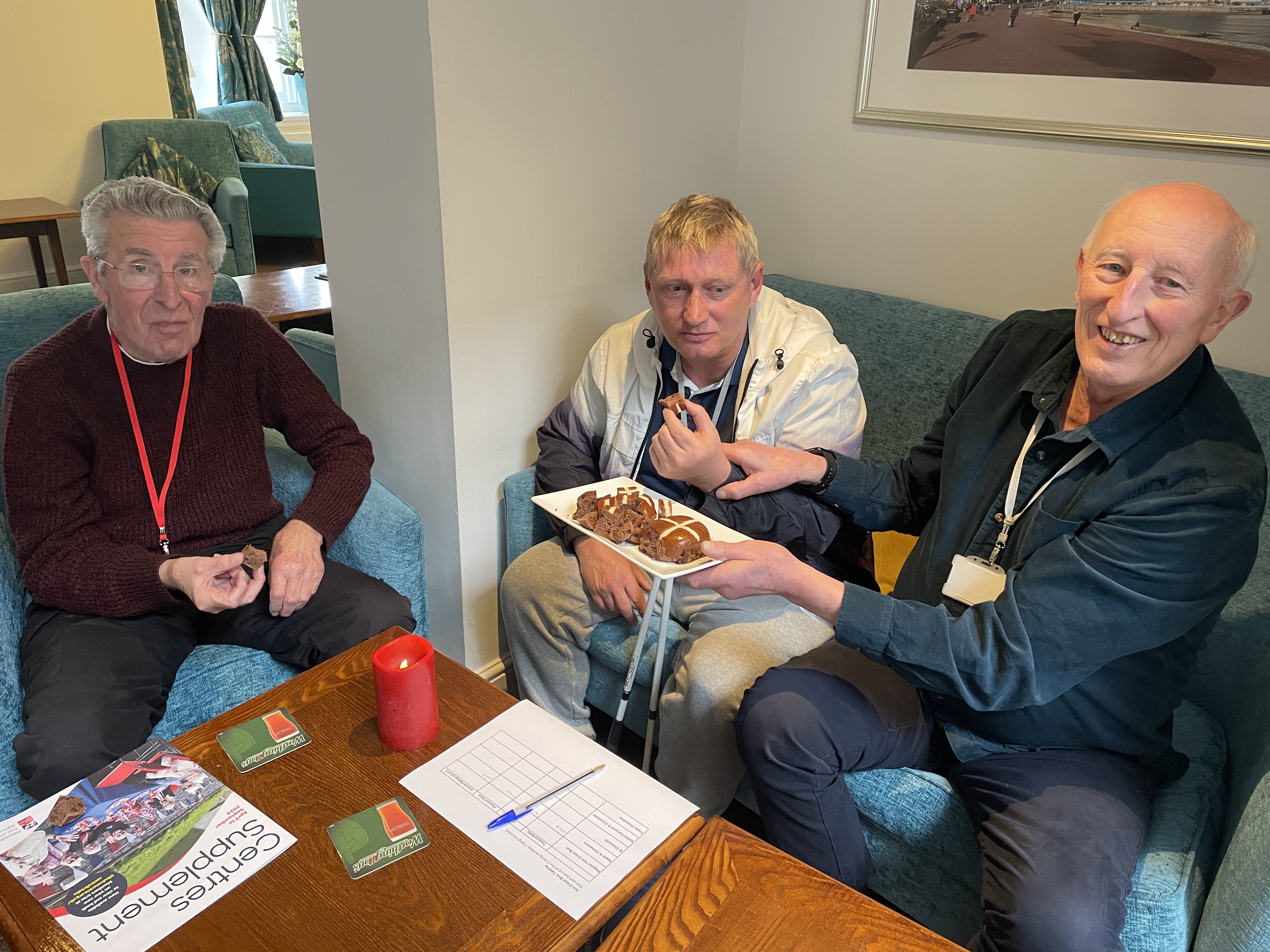 Three men sit at a table, two are holding pieces of hot cross bun, while the third holds a plate of hot cross bun pieces.
