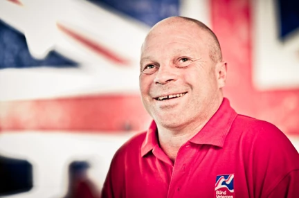 Blind veteran Billy smiling, wearing a red Blind Veterans UK polo shirt, with a British flag design covering the wall behind him.