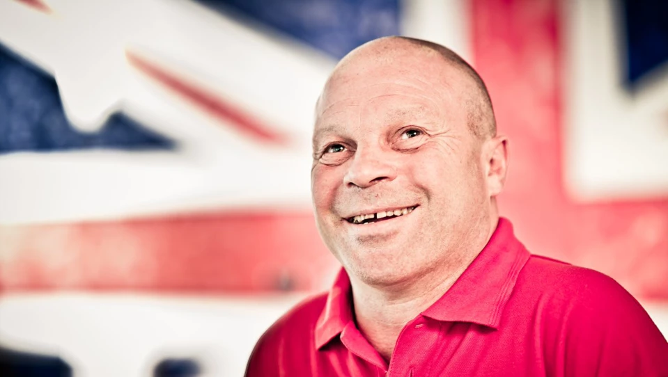 Blind veteran Billy smiling, wearing a red Blind Veterans UK polo shirt, with a British flag design covering the wall behind him.