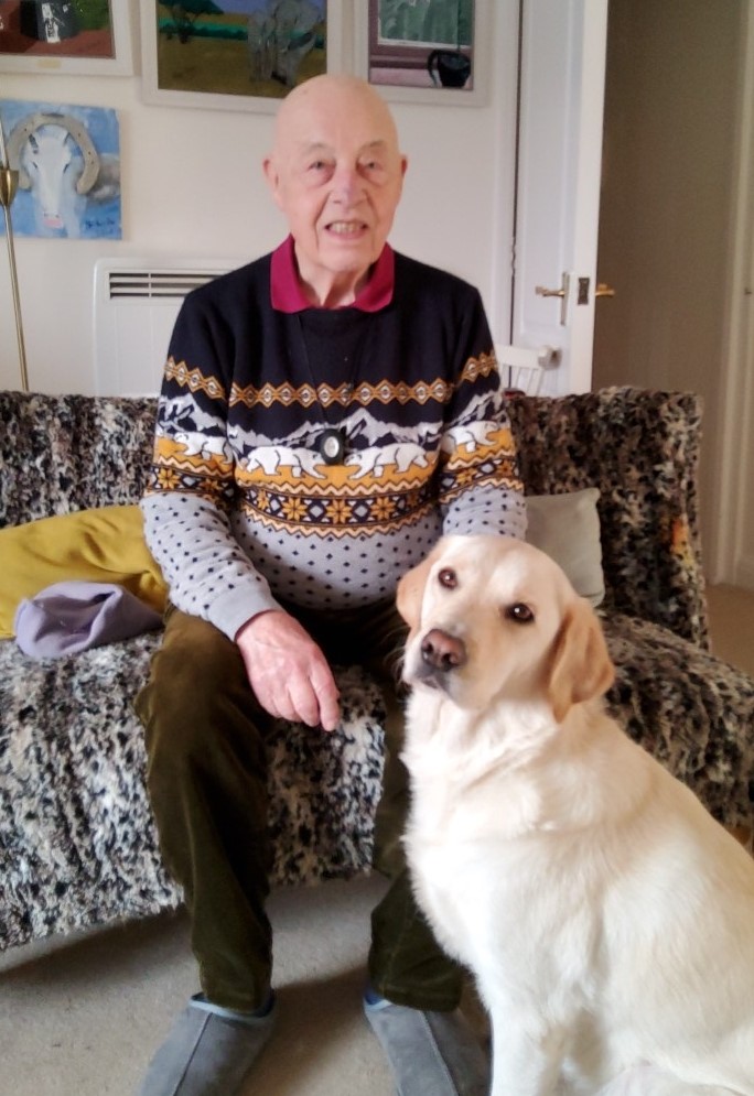 Tom is wearing a Christmas jumper and is sitting on the sofa with his guide dog at his feet