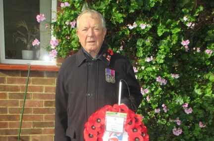 Blind Veteran Jim stood in his garden wearing a poppy and displaying his medals. He is holding his white cane and his poppy wreath.
