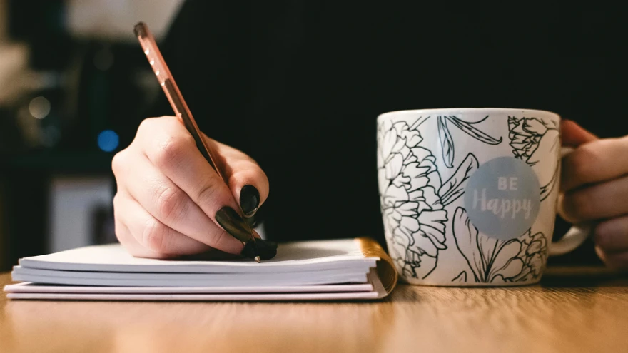 A photo of a person writing on paper while holding white mug with flowers and the words 'be happy' painted on it