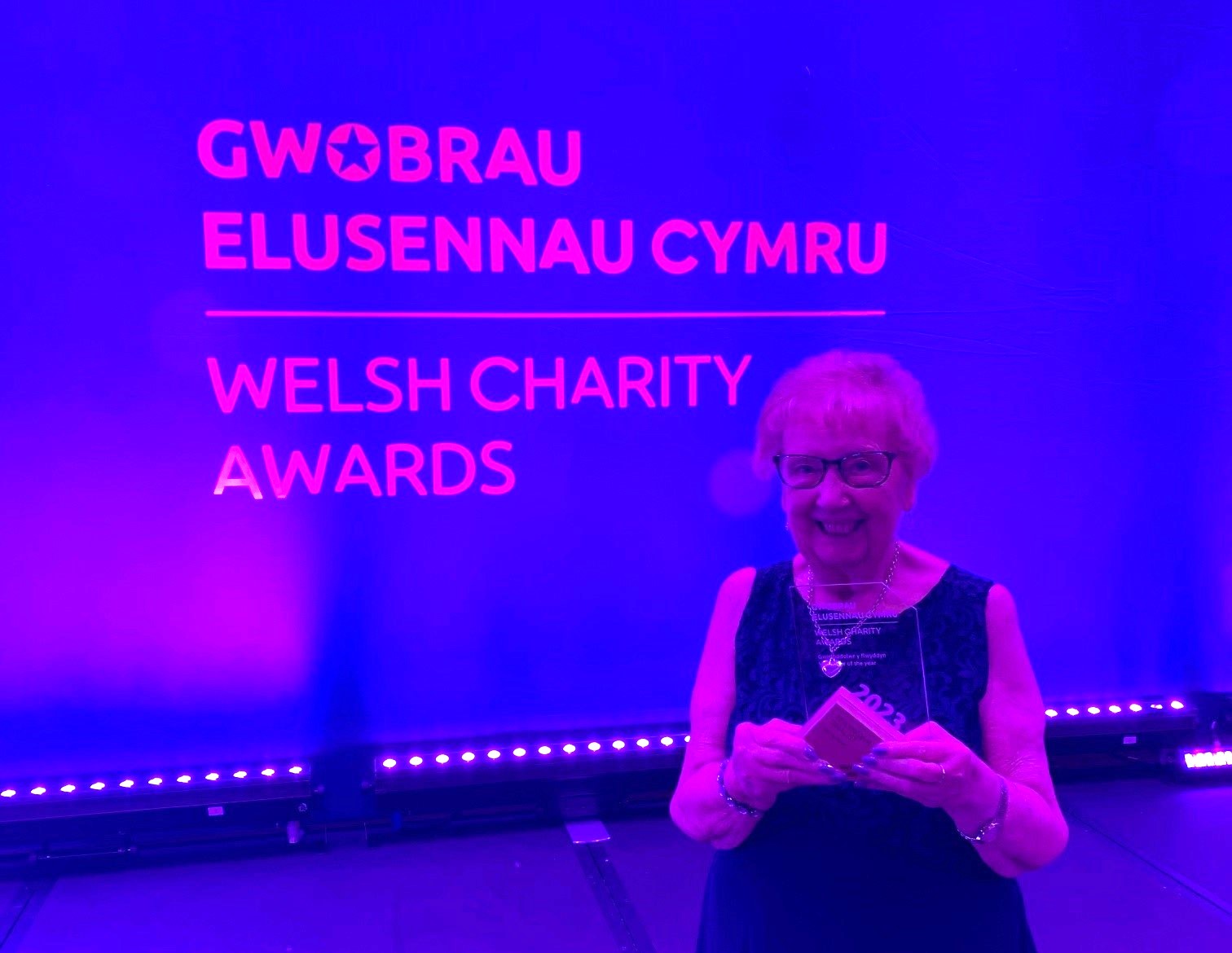 Gill stands in front of a branded screen and holds her award proudly while smiling