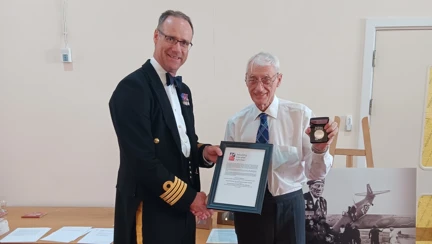HMS Sultan Commanding Officer wearing officer mess dress shakes the hand of James while presenting a commemorative coin. Behind them is a black and white image of D-Day veteran Alec.
