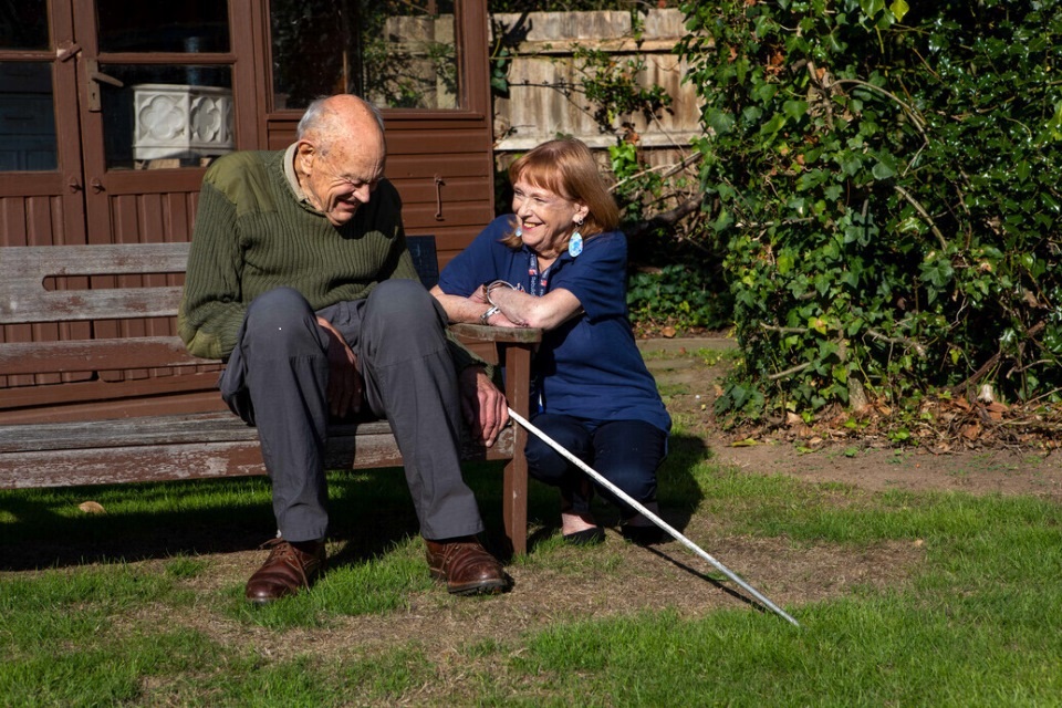 Mike sits on a bench and Jennie crouches next to him on a beautiful sunny day