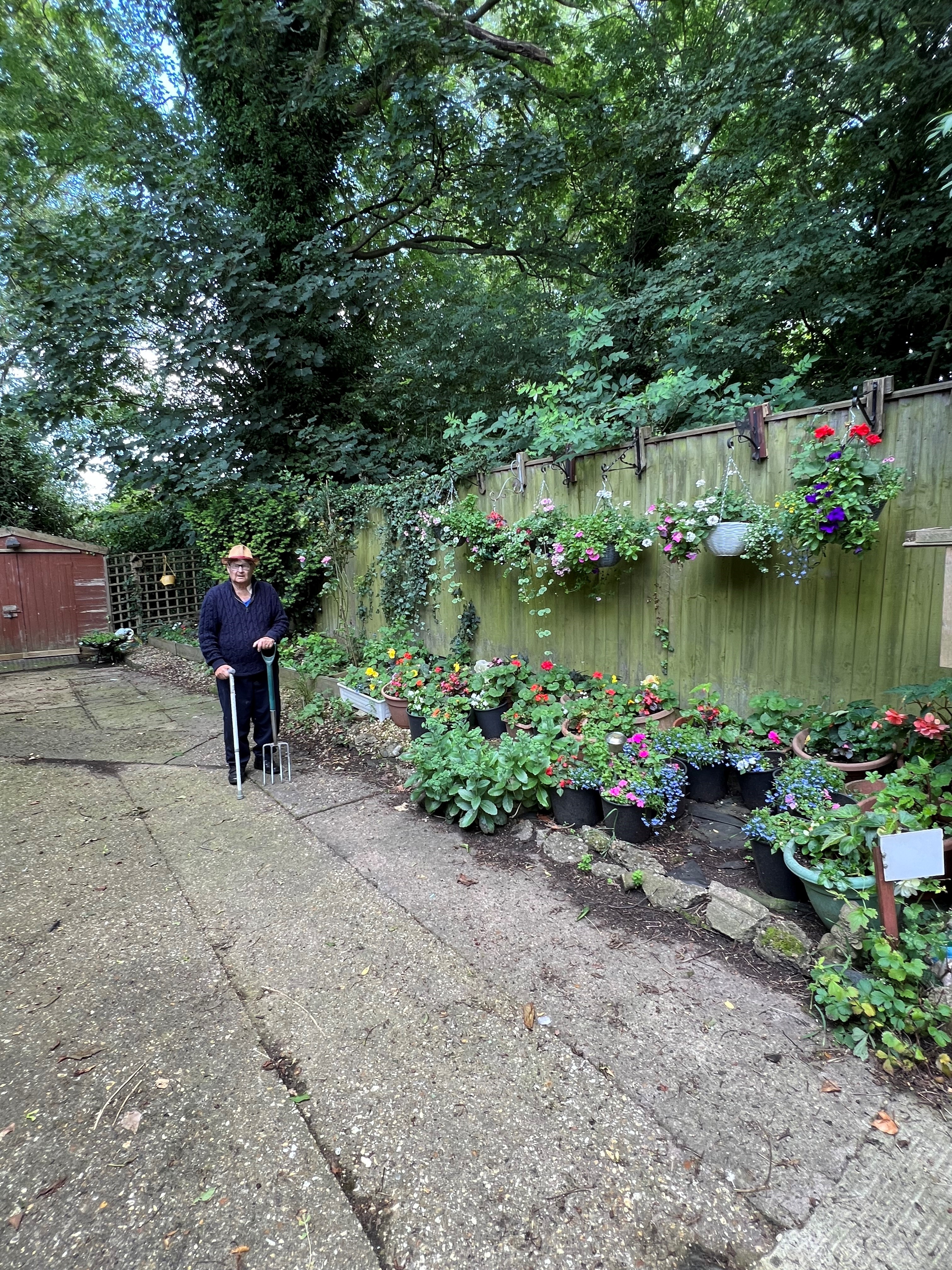 Lawrence stood with a walking stick in one hand a garden fork in the other. His garden shed is behind him and to his left are lots of pots and hanging baskets in bloom with red, yellow, blue, pink and white flowers