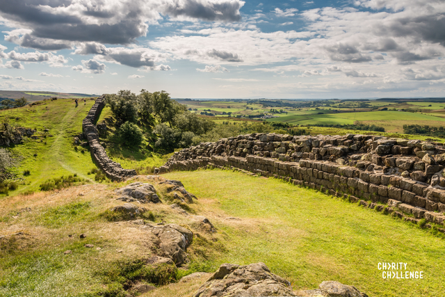 The remains of the stone wall stretching off into the distance across beautiful green landscape. The sky above is blue with dramatic grey and white clouds.