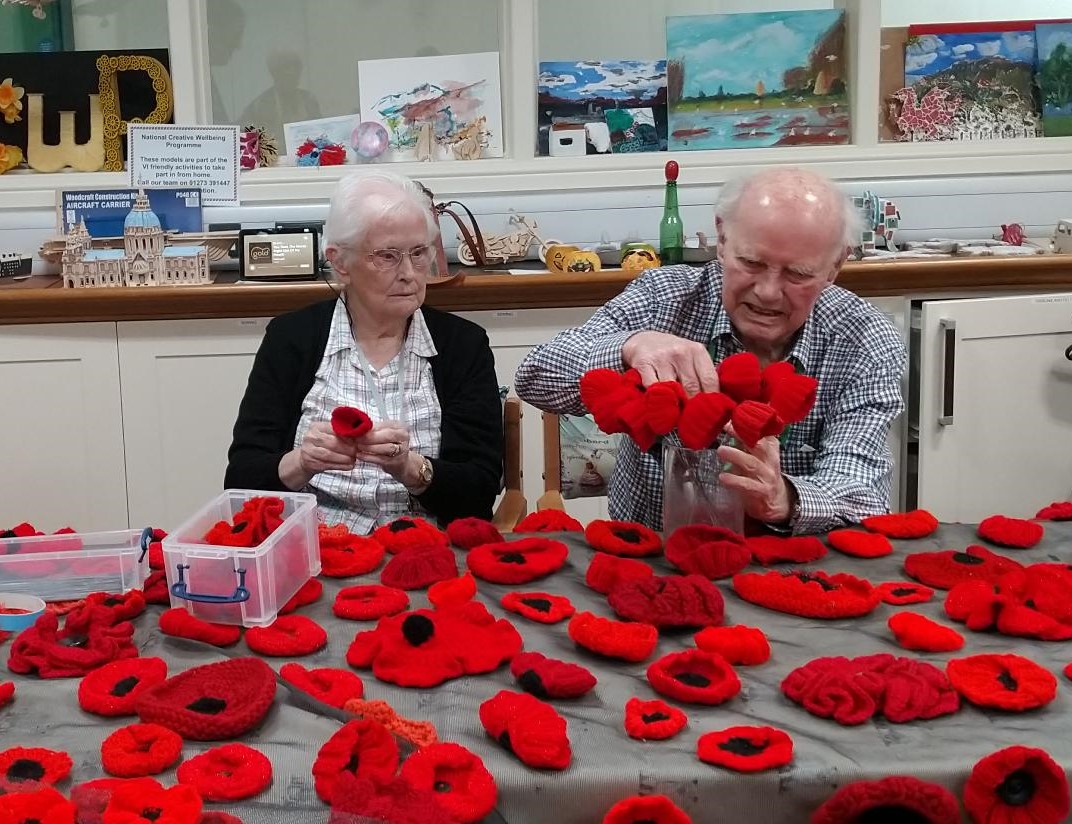 George and Shirley are sat at a table full of knitted poppies in the craft room at the Llandudno Centre