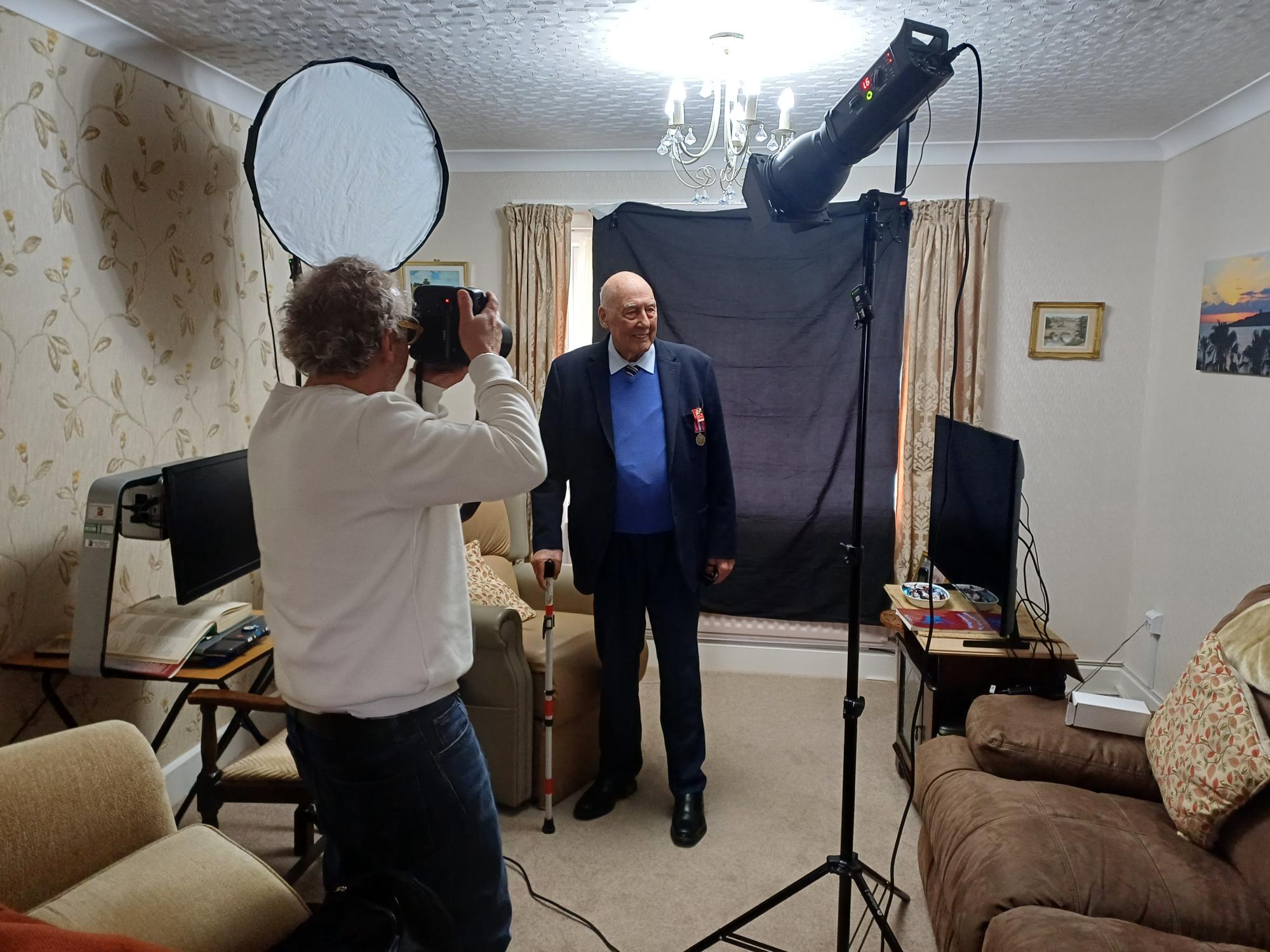 Arthur stands for a photograph in his living room as Richard Cannon holds a large professional camera. In the background, there is a black sheet background and a lighting set up.