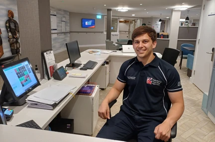 Jacob, dressed in a navy Blind Veterans UK polo shirt, sits at a desk in the Rustington Centre of Wellbeing. The background shows a modern office environment with computer monitors, paper work and hallways leading to other areas at the centre.