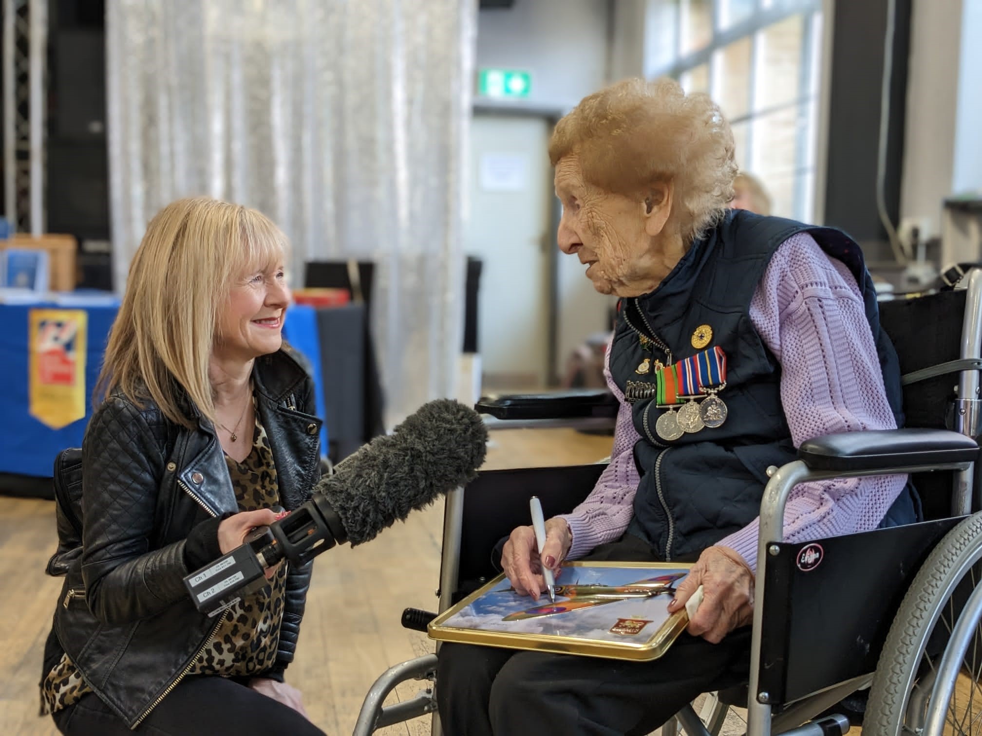 Margaret is wearing her medals and speaking into a microphone held by a journalist from the BBC