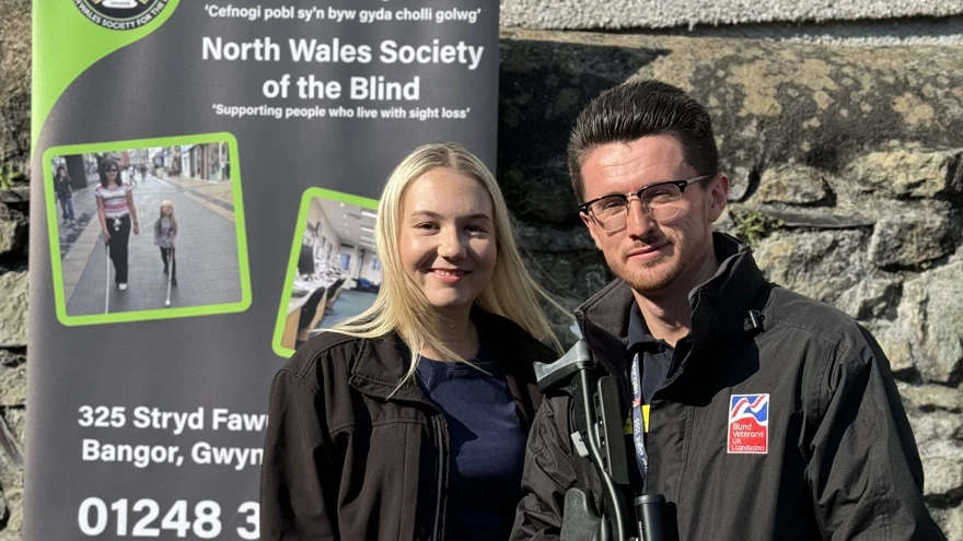 A lady with blonde hair stands next to a man with brown hair. The man is holding an air rifle and there is a banner in the background with the words North Wales Society of the Blind