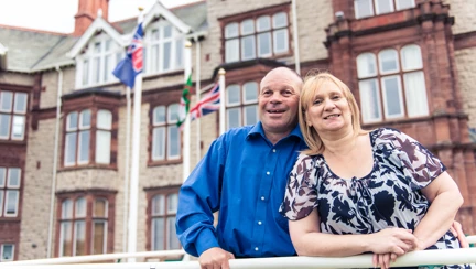 Blind veteran Billy with his arm around his wife, smiling as they stand outside our Centre of wellbeing in Llandudno