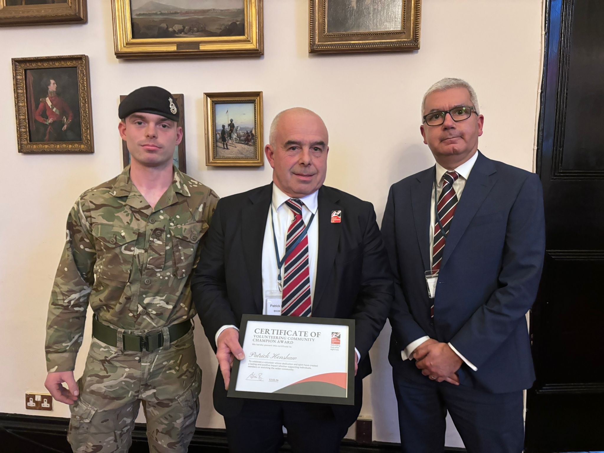 Volunteer Patrick holding his Founder’s Award certificate flanked by his son Matthew in British Army uniform and Salman Shamim of Corps Security at RMA Sandhurst