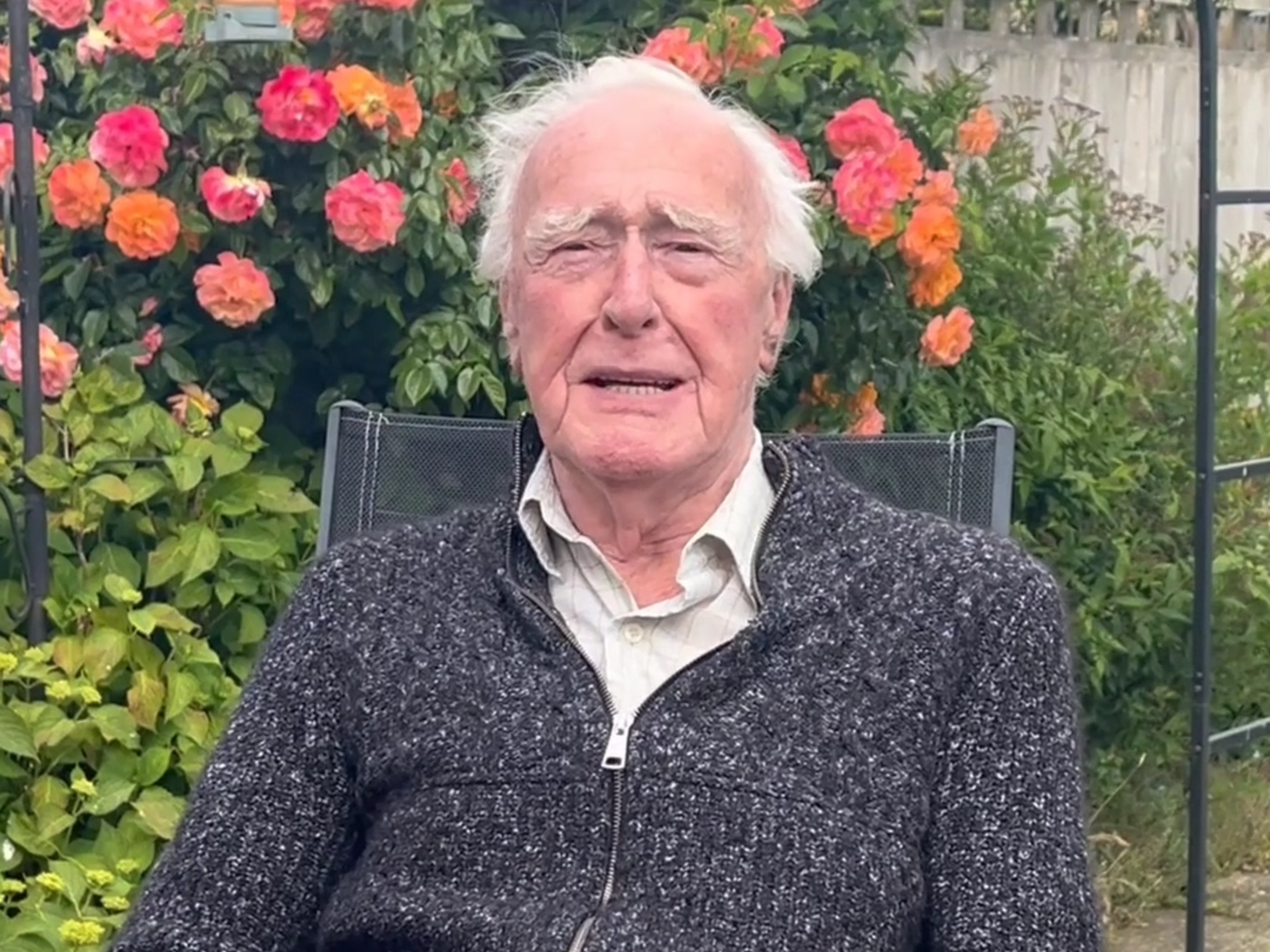 Blind veteran Colin sits on a chair outside in his garden, shrubbery and flowers are seen in the background. He is mid speech.