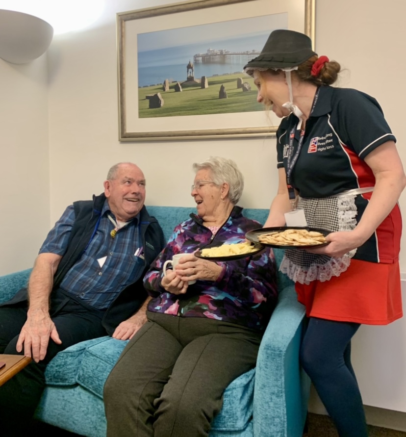 A man and woman sit on a sofa being served cheese by another lady wearing traditional Welsh dress.