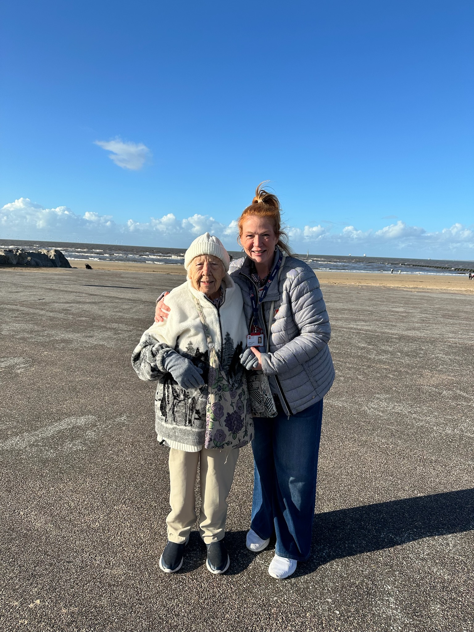 Blind veteran Olly and volunteer Lanie smiling and standing arm-in-arm on a breezy beach promenade in New Brighton.