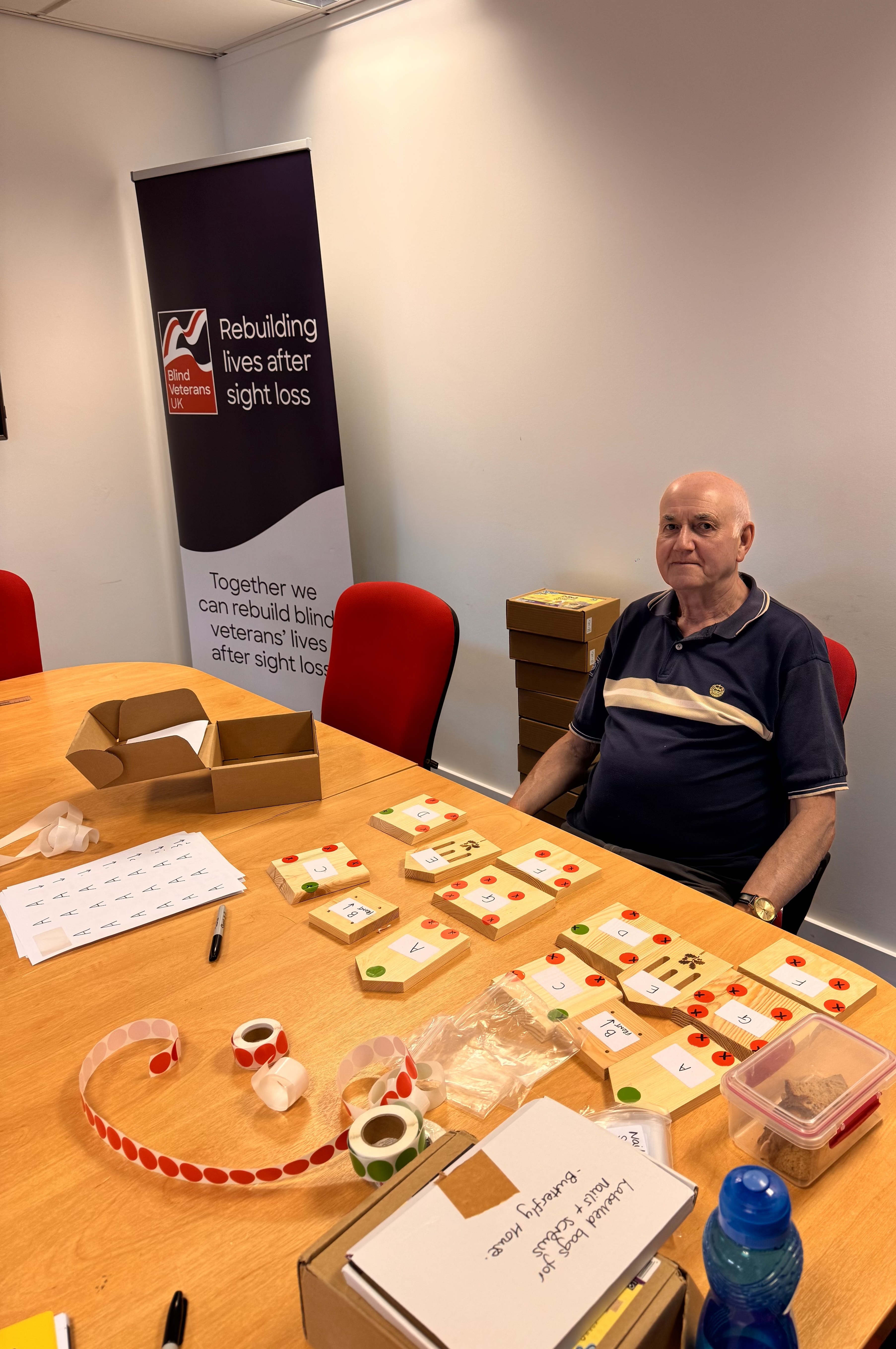 Martin, a volunteer, sits at a table neatly covered in organised craft materials and butterfly house kit components. Behind him is a Blind Veterans UK banner reading “Rebuilding lives after sight loss.”