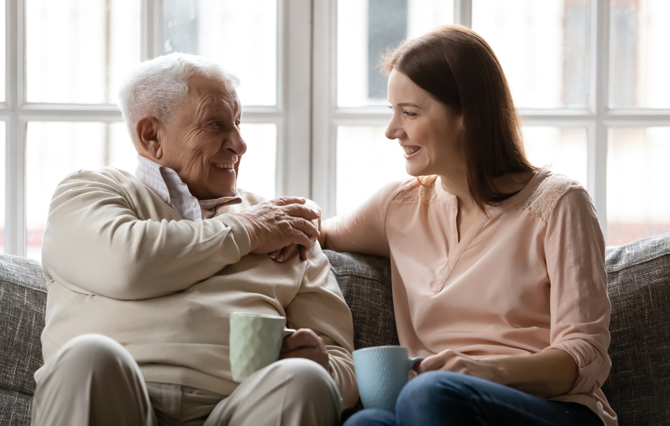 A stock photo of an Elderly man smiling while talking to a younger woman whilist having a cup of tea