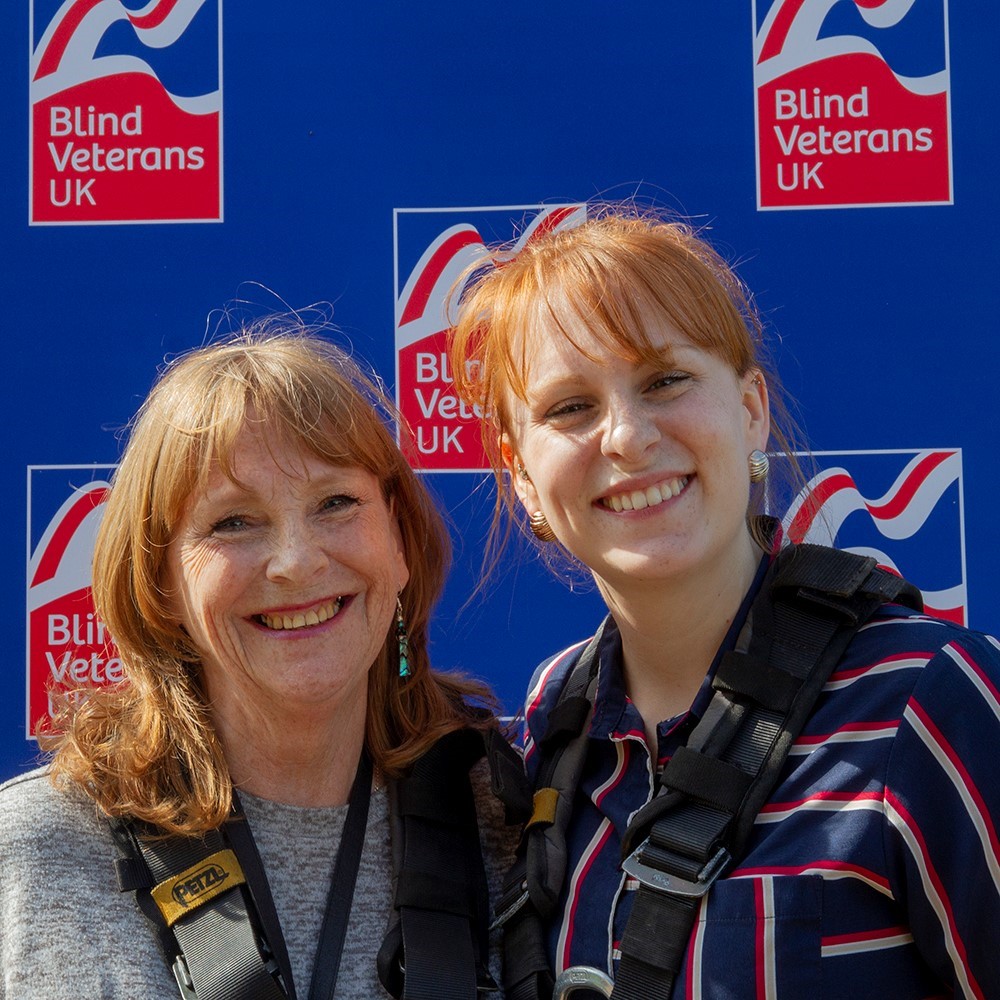 Jennie and Fern are both wearing a harness and standing in front of a Blind Veterans UK branded wall