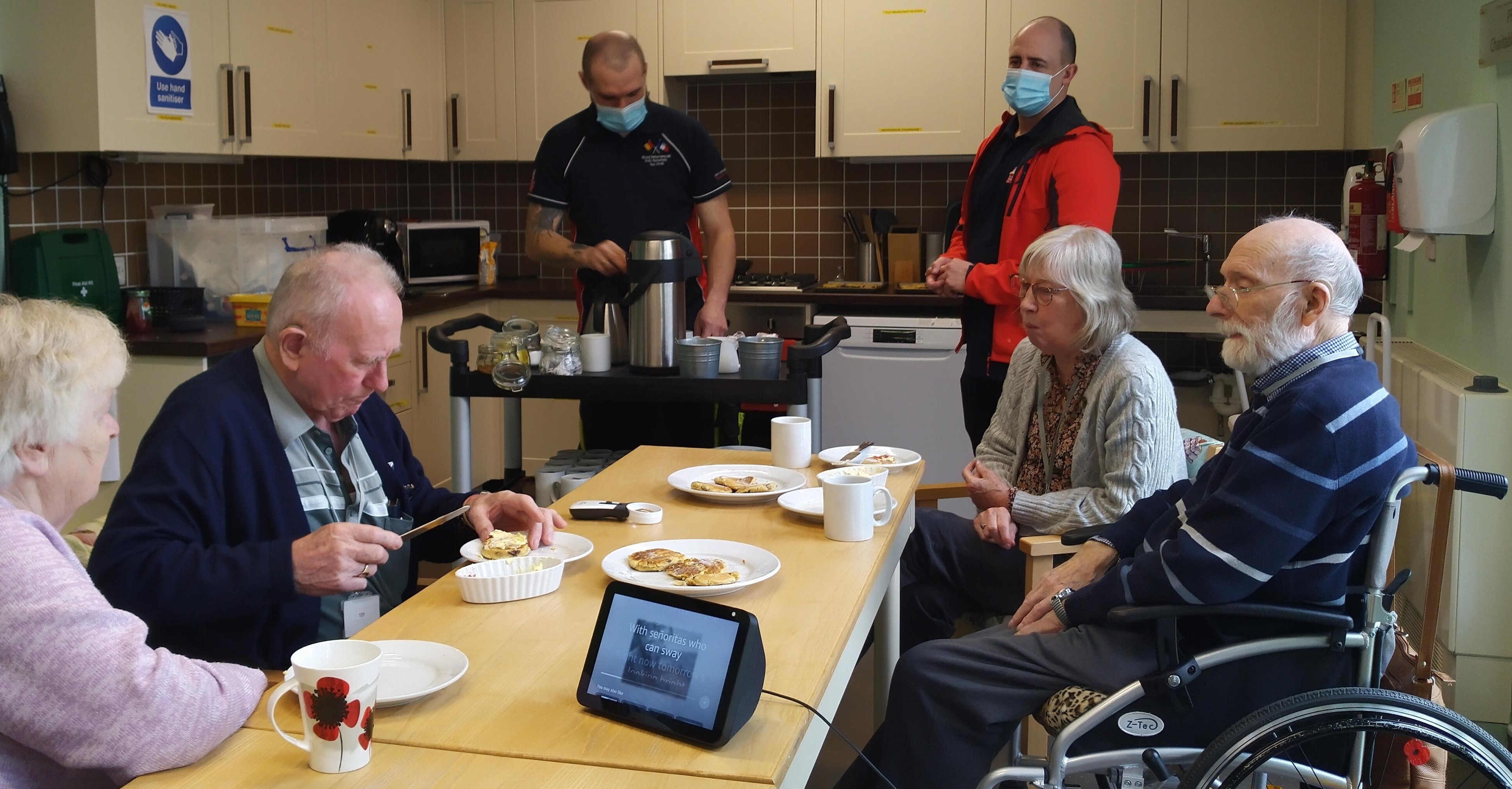 A photo showing a group of blind veterans and their wives enjoying a cup of tea with the scones they baked, assisted by Alexa