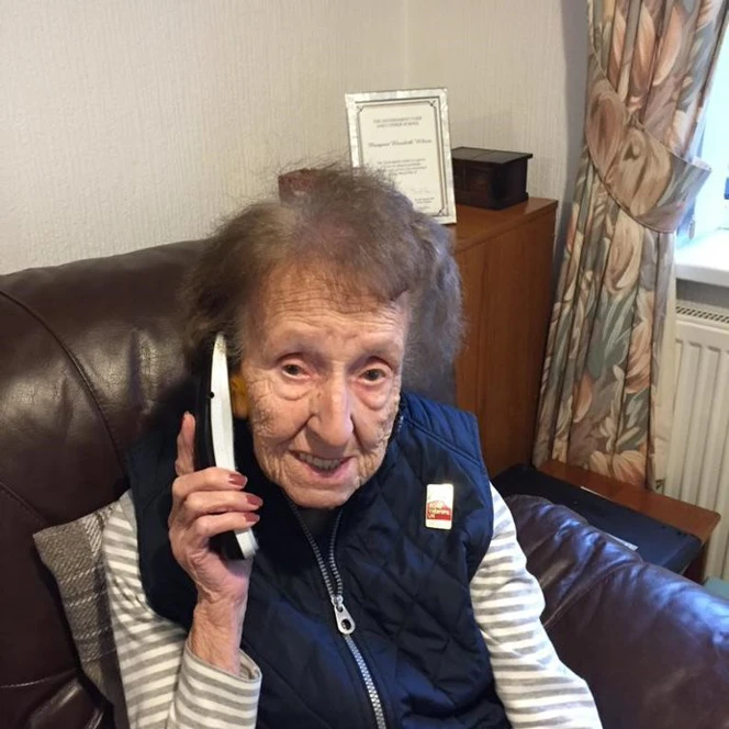 Blind veteran Margaret, sitting on an armchair, smiling to the camera as she holds a phone up to her ear