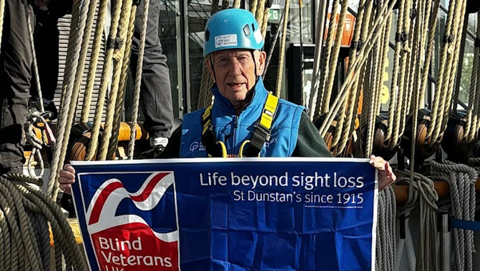 Kelso wearing a helmet and harness is stood onboard the Cutty Sark ahead of his climb holding a Blind Veterans UK banner