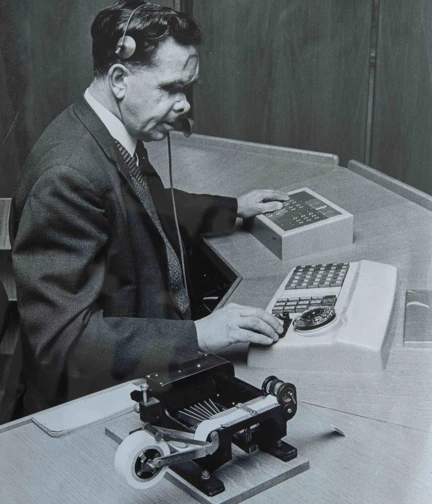 A black and white photograph of a vision-impaired veteran operation a telephone switchboard. He is wearing a headset and using a Braille notetaking device.