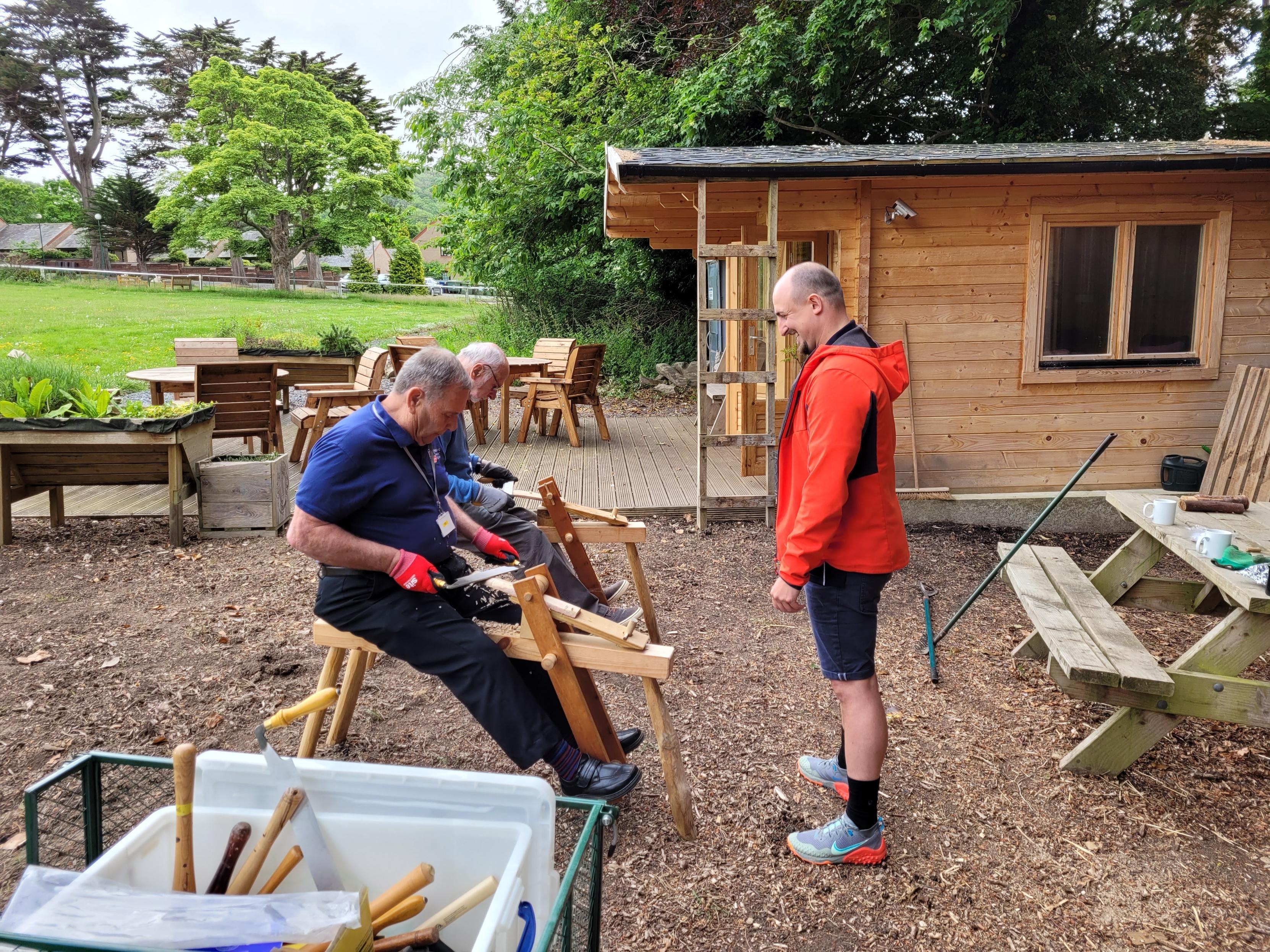Two blind veterans woodworking, with a wooden cabin in the background