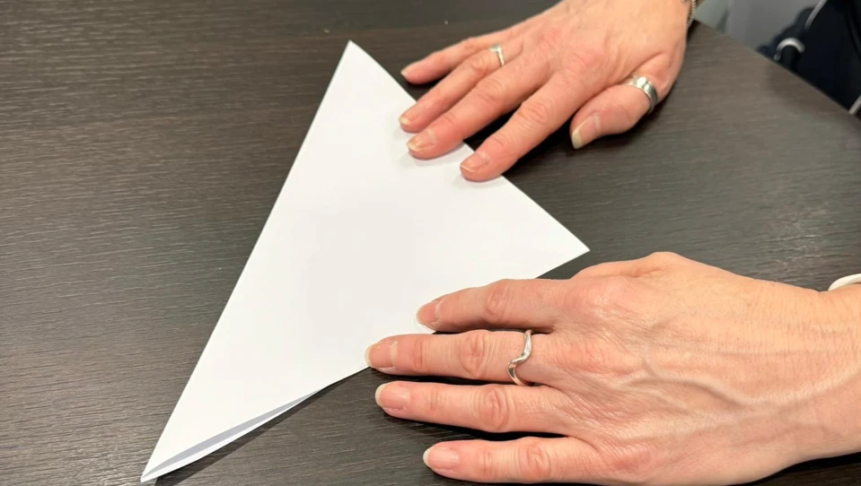 A person’s hands resting on a dark table. They are folding a piece of paper diagonally into a triangle, which is the first step in creating a paper snowflake. One hand is holding the paper steady, while the other is positioned nearby, ready to assist with the fold.