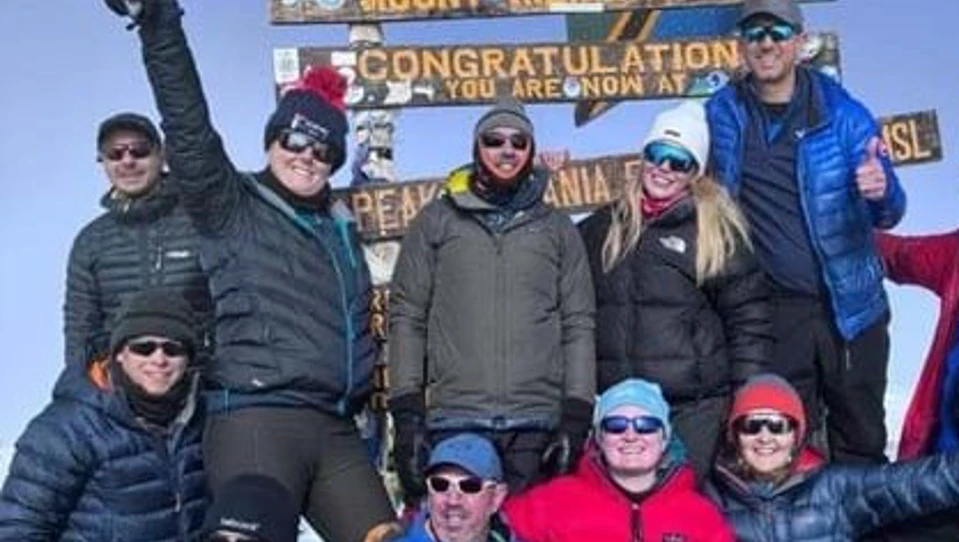 Group of smiling people bundles up in warm coats, gathered together under a sign that reads 'Mount Kilimanjaro. Congratulations. You are now at the peak'