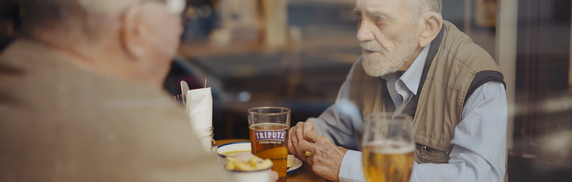 Blind veteran Ken and volunteer Liam sitting down, chatting in a pub, with drinks and food on the table