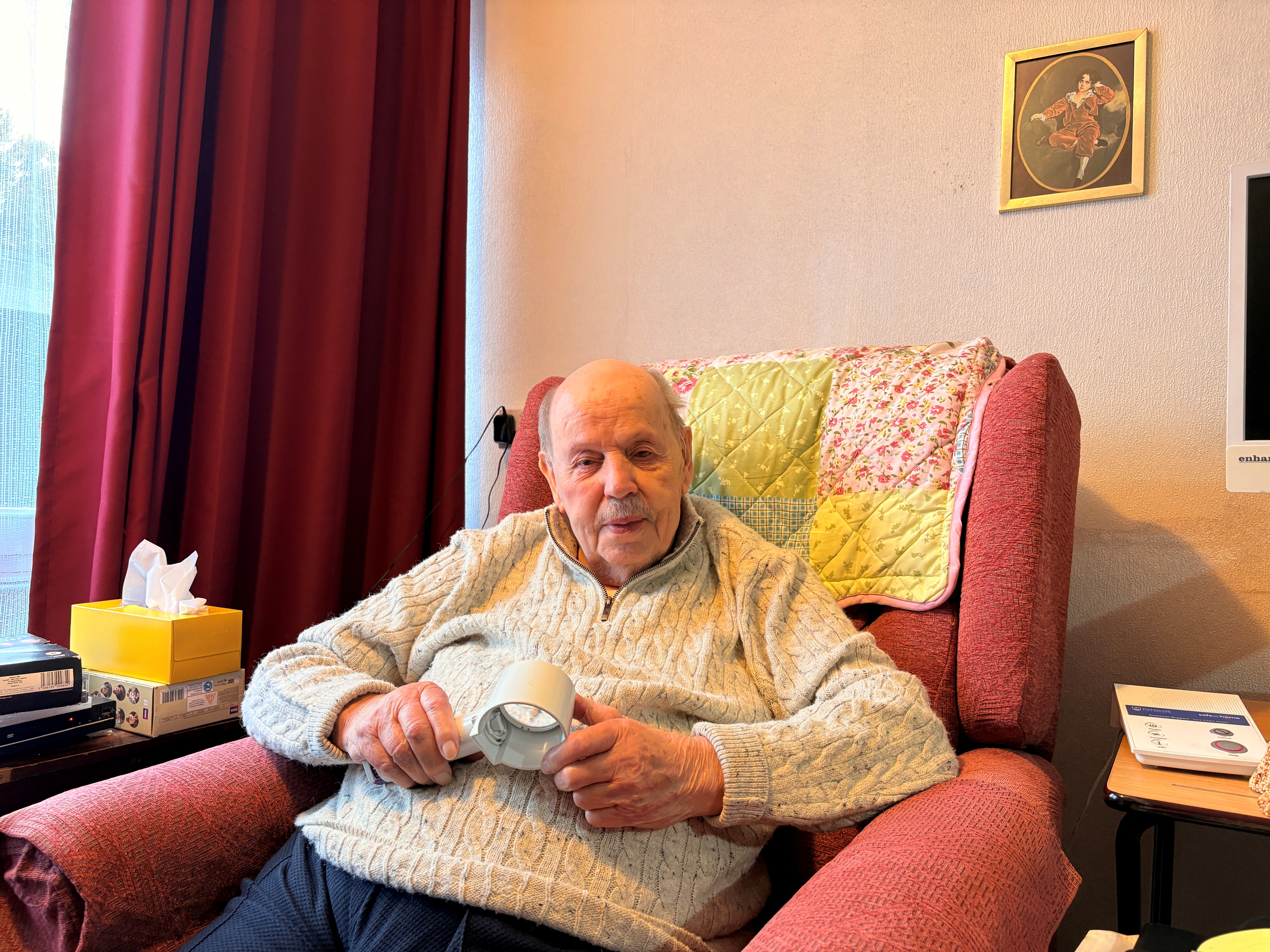 Doug sits at home and looks into the camera, he is wearing a winter jumper and holding a hand magnifier