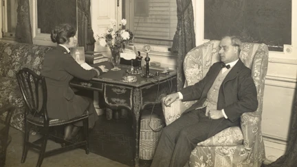 A black and white photograph of Sir Arthur Pearson seated in an armchair in his office at St Dunstan's Lodge. He is dressed in a suit and appears deep in though. A woman sits at a nearby desk writing.