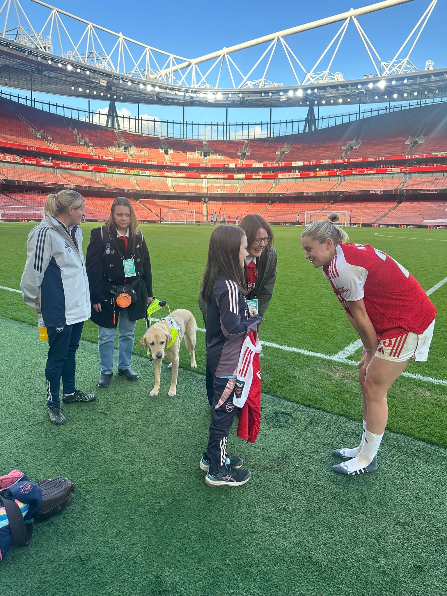 Stood on the edge of the pitch at the Emirates Stadium, Kelly and her guide dog Archie talk to Arsenal hero Kelly Smith while player Alessia Russo bends down to speak with 10-year-old Bethany who is holding the signed shirt she's been presented with. Sarah is stood next to Bethany watching on and smiling.