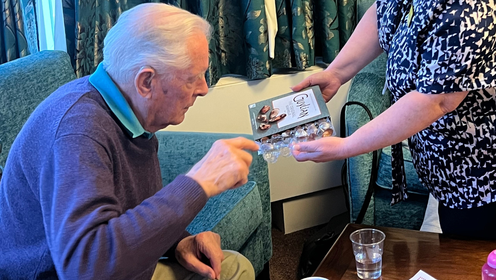 A blind veteran reaching into a box of chocolates as he sits comfortably on a sofa
