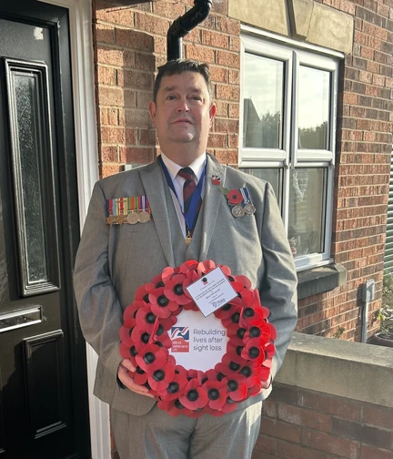 Clive is wearing a grey suit and is stood in front of his front door holding his wreath