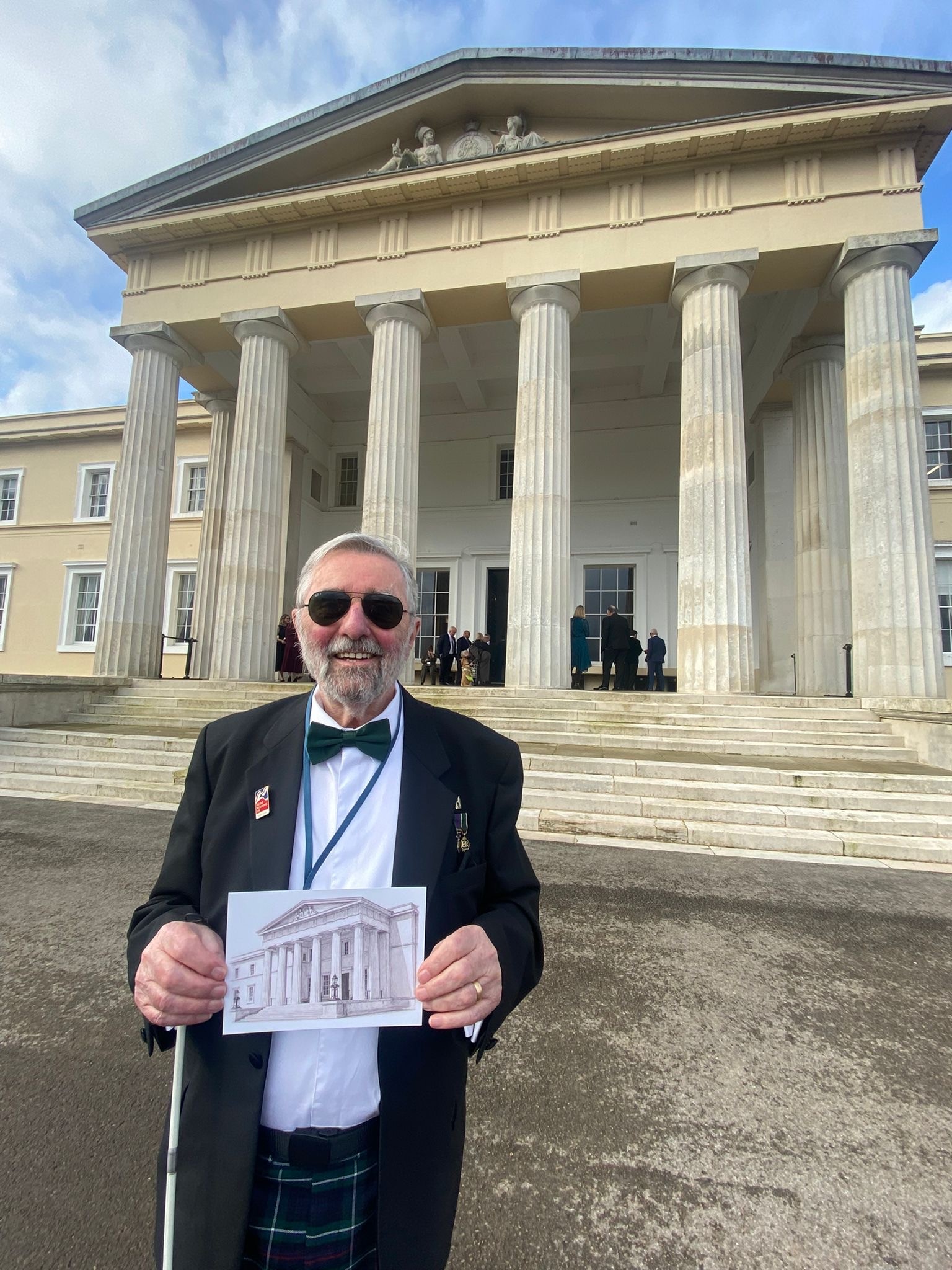 Blind veteran Bryan standing on the steps of Old College at RMA Sandhurst in formal attire with bow tie and dark glasses, holding up his line drawing of the same building with the actual classical columned facade visible behind him.
