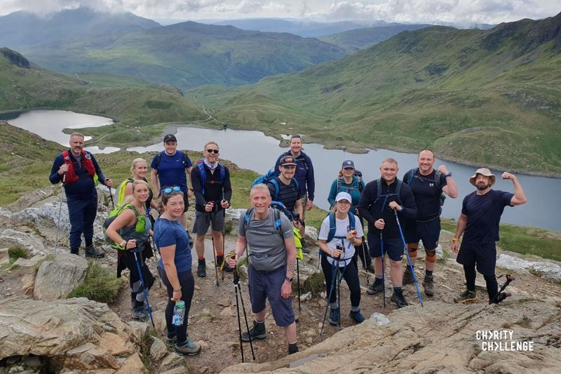 A group of challengers on a rocky hill side with mountains, lakes and blue skies behind them. They are smiling and some are making 'strong man' poses - flexing their biceps!