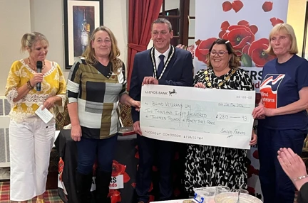 A group photo of five people including blind veteran Jill in a Blind Veterans UK t-shirt. The group is holding an oversized cheque