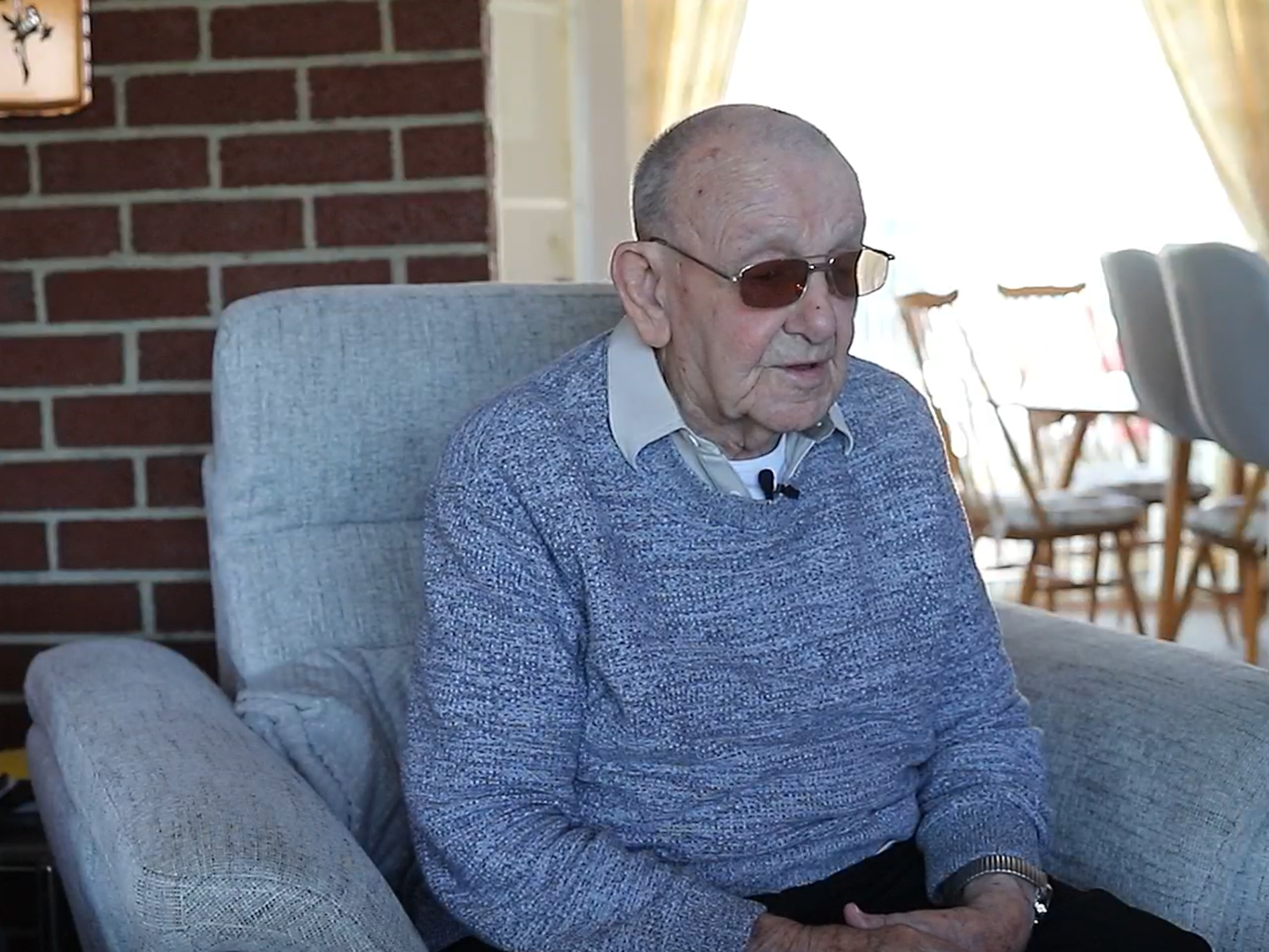 Ken is wearing a blue jumper and dark glasses as he sits in a comfortable armchair at home, speaking about his wartime memories.